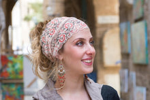 A woman wearing a beige lace headband with red and green floral embroidery, styled in a half-up bun, shown in a softly blurred outdoor setting.