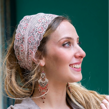 A woman in profile wearing a beige lace headscarf with red and green floral embroidery, styled with dangling red and gold earrings against a dark green door and stone wall background.