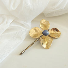 Gold flower-shaped brooch with a central blue gemstone on a white fabric background

