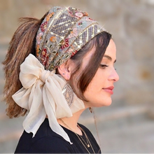 Woman wearing a decorative headscarf with a bow, standing outdoors.


