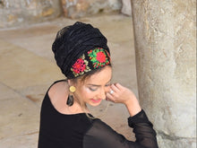 A woman wearing a black lace headwrap turban with vibrant red and green floral embroidery, paired with a black long-sleeve top and gold earrings, sitting next to a stone pillar.