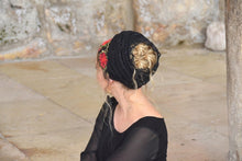 A woman shown from the back wearing a black headwrap or hair accessory with red floral embroidery, paired with a black top, set against a rustic stone wall background.