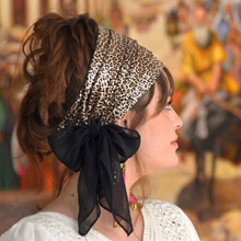 Woman wearing a leopard print headscarf with a black bow, standing in front of a blurred background.

