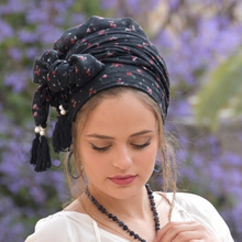 Woman wearing a black patterned headscarf with tassels against a blurred purple floral background

